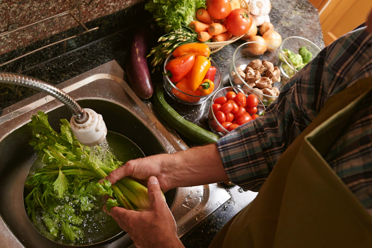 A man washing celery in a kitchen sink with lots of vegetables next to him.
