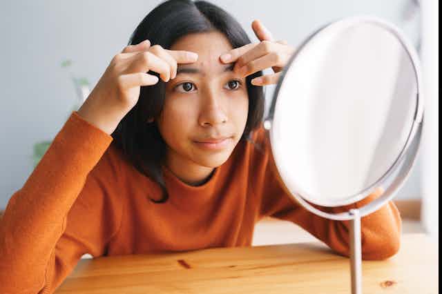 A young woman examines acne on her forehead in a mirror.