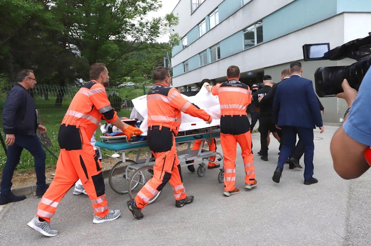 Os paramédicos levam uma maca até um hospital, segurando lençóis para cobrir a pessoa na maca.
