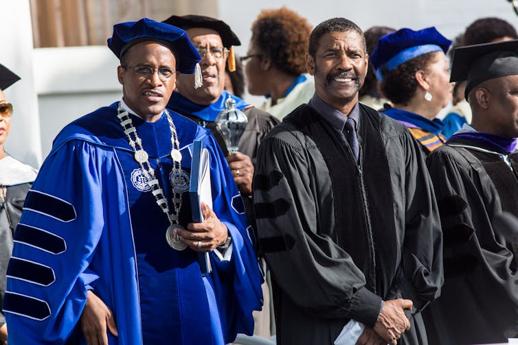 Two men in graduation gowns – one blue and one black – stand next to each other.