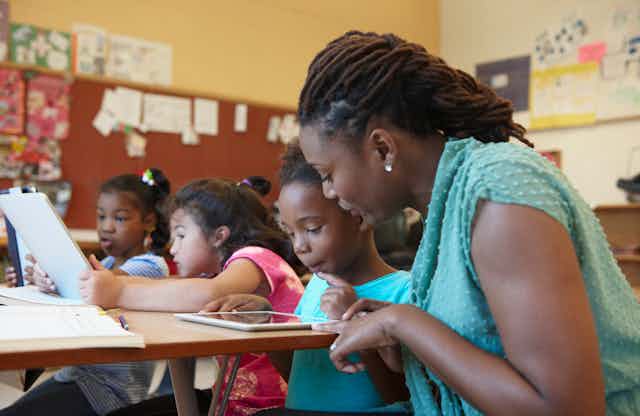 A teacher helps a student on a tablet at a desk.