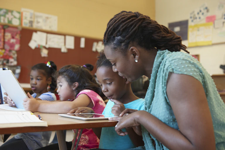 A teacher helps a student on a tablet at a desk.