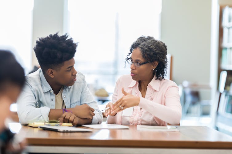 A female teacher helps a male student at his desk.