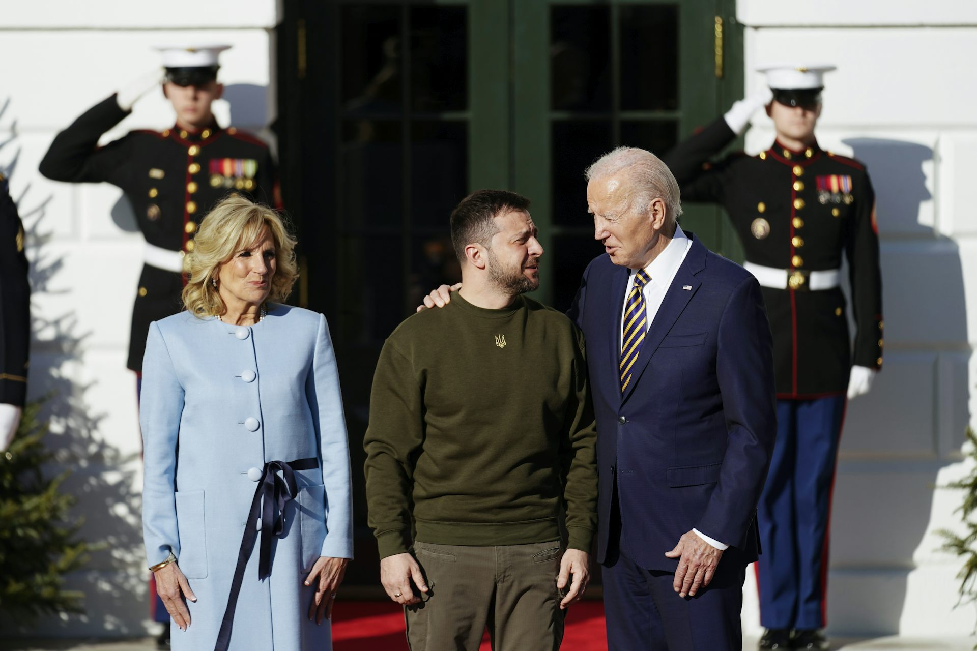An older man in a suit puts his arm around a dark-haired man dress in dark green casual clothing. A blond woman in a pale-blue suit smiles beside them and guards salute behind them.