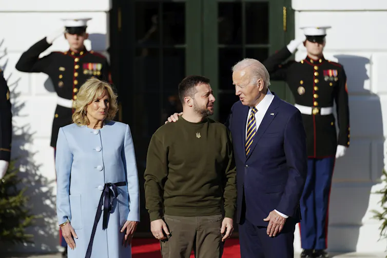 An older man in a suit puts his arm around a dark-haired man dress in dark green casual clothing. A blond woman in a pale-blue suit smiles beside them and guards salute behind them.