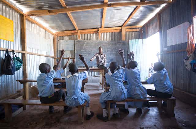 A small group of schoolchildren in light blue school uniforms raising their hands to address a teacher sitting on a  desk at the head of the class