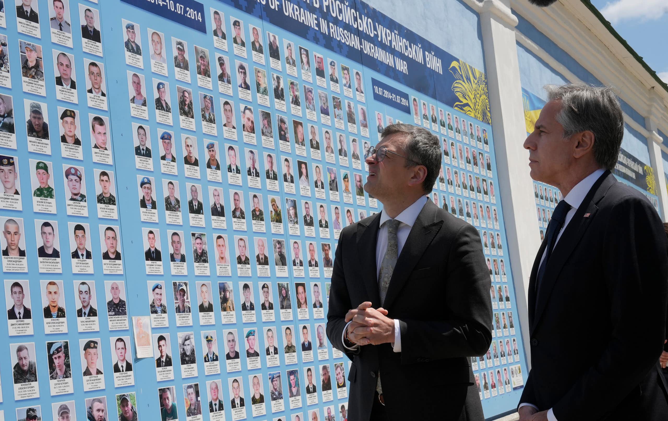 Two men in suits look at a large mural features the faces of fallen soldiers.