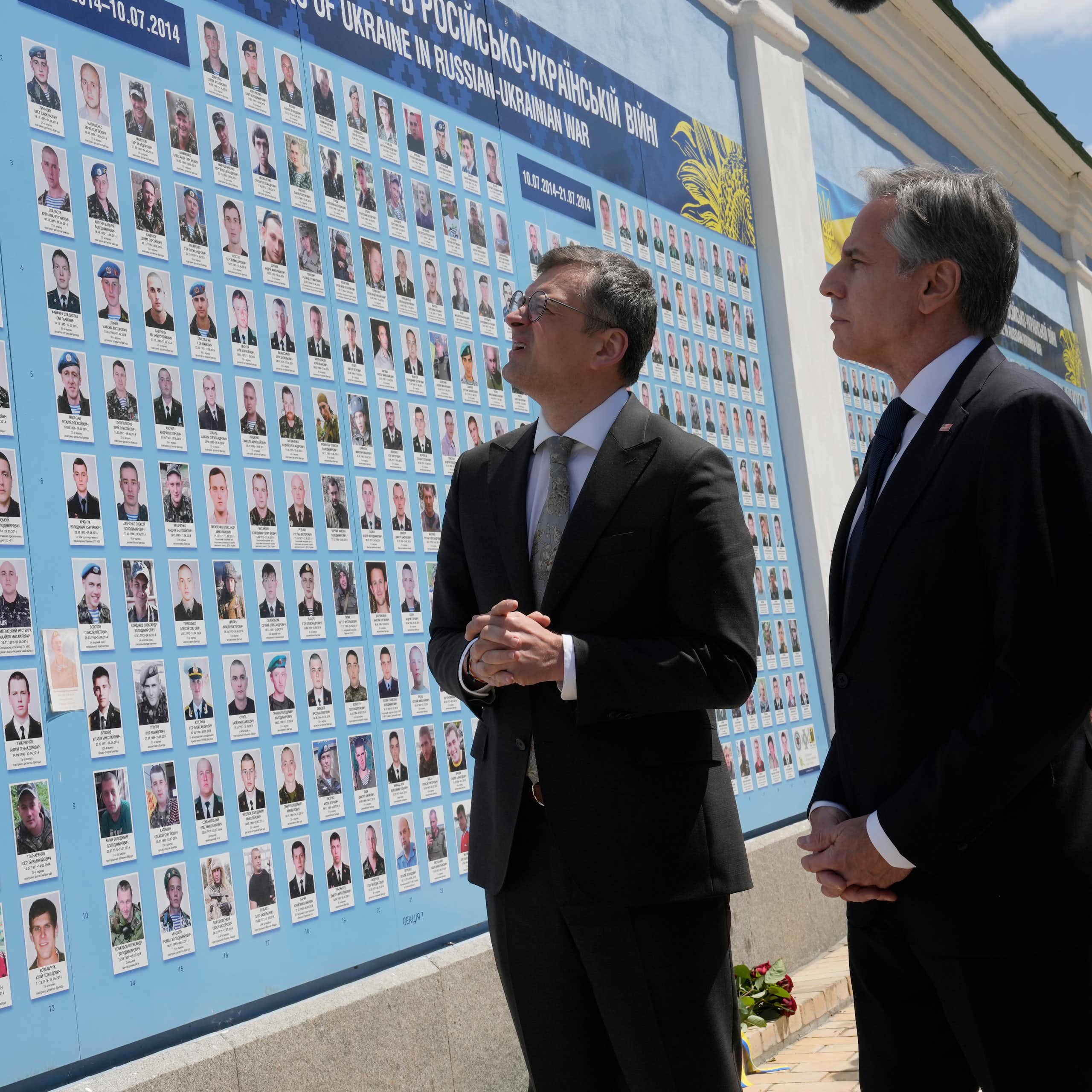 Two men in suits look at a large mural features the faces of fallen soldiers.