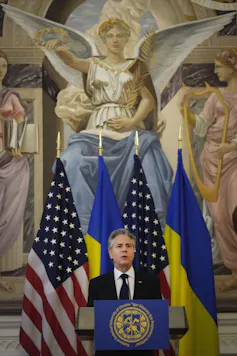 A man speaks behind a lectern with flags and an ornate painting behind him.