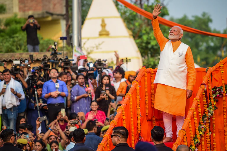 A man in an orange tunic waves as in the background TV journalists watch on.