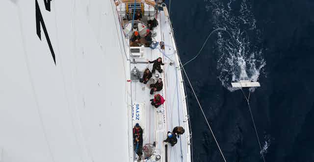 Shot from top of mast looking down onto sailboat, sea to right with trawling white equipment