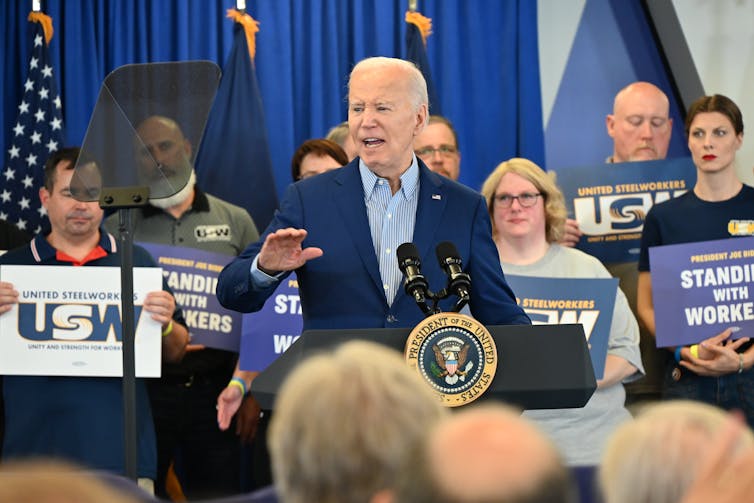 Biden speaks at a podium, with people behind him holding United Steelworkers signs.