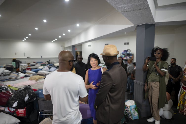 A woman in a suit seen speaking with people in a room with cots.