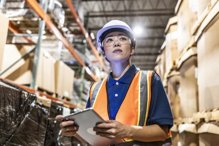 A warehouse employee wearing an orange safety vest is seen taking inventory with digital tablet.