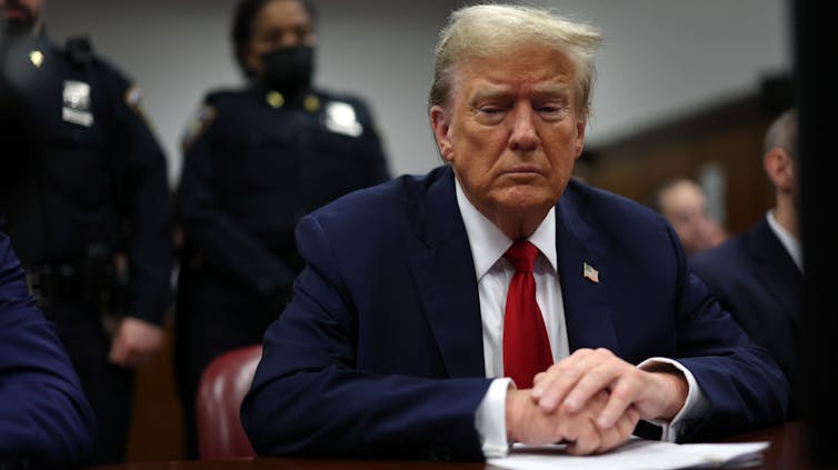 A man in a blue suit jacket, red tie and with hands folded, sitting at a table with police behind him.