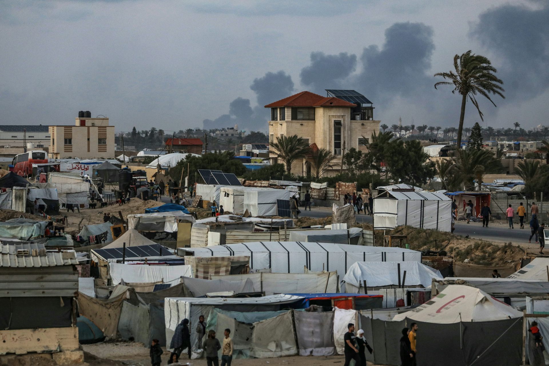 A makeshift refugee camp with a large plume billowing in the distance.