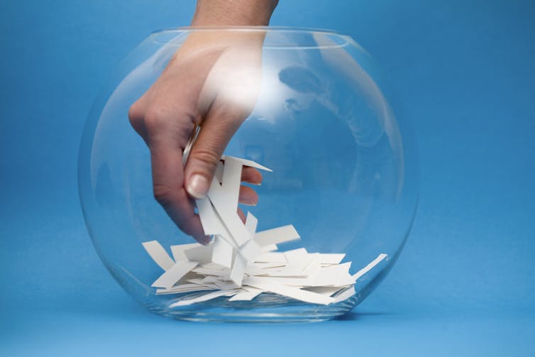 A hand reaches into a glass bowl to select a piece of paper from a group of pieces of paper.