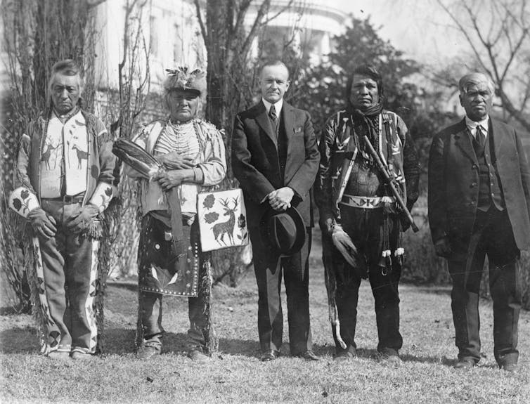A black and white photo of five men standing formally outside, with two in suits and three in Native American clothing.