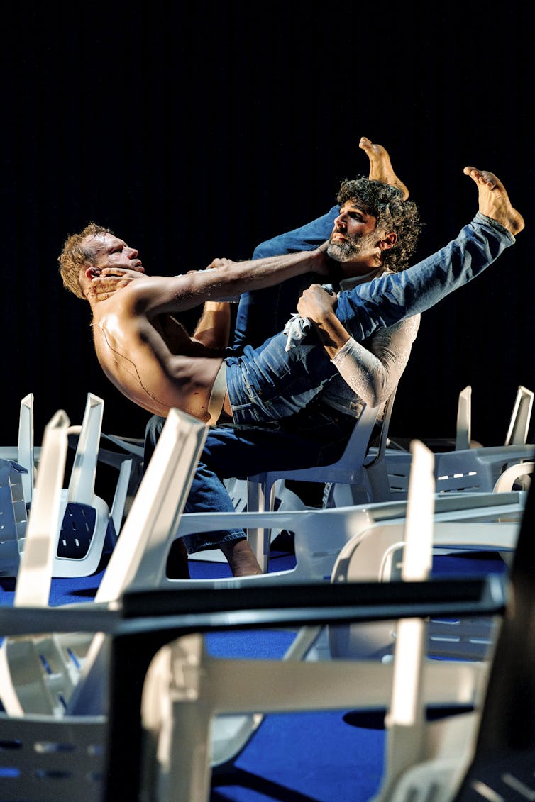 Two men could perhaps be wrestling above white plastic chairs.