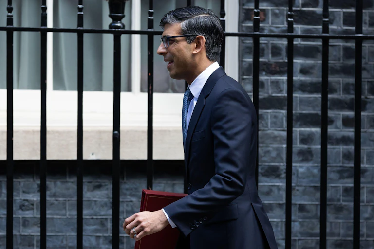 Side view of prime minister Rishi Sunak walking in front of Downing Street, holding a red ministerial folder under his arm