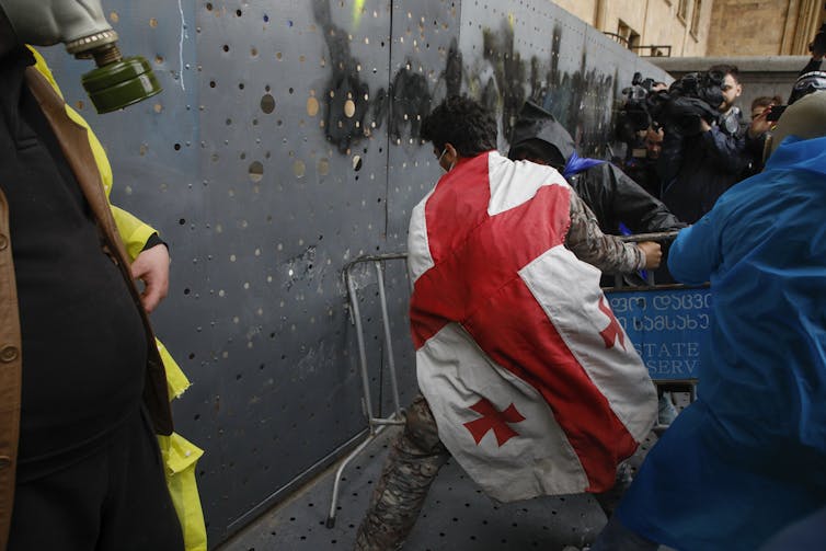A protestor draped in the Georgian flag attacking a barrier protecting Georgia's Parliament building.