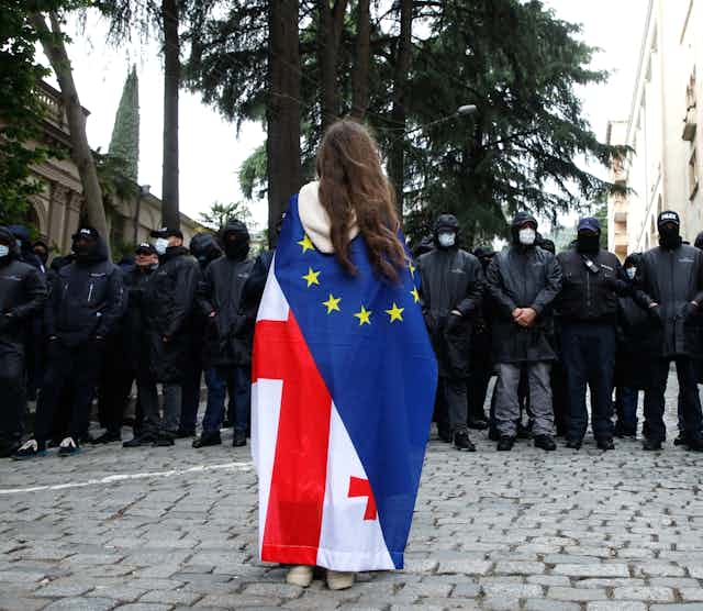 A protester wearing a Georgian and European flag faces off with policemen blocking a street.