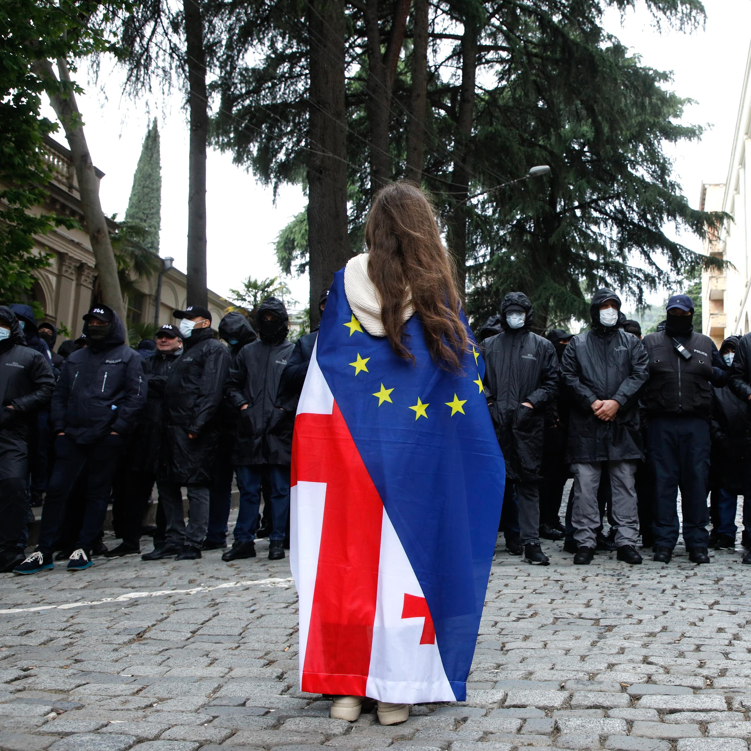 A protester wearing a Georgian and European flag faces off with policemen blocking a street.