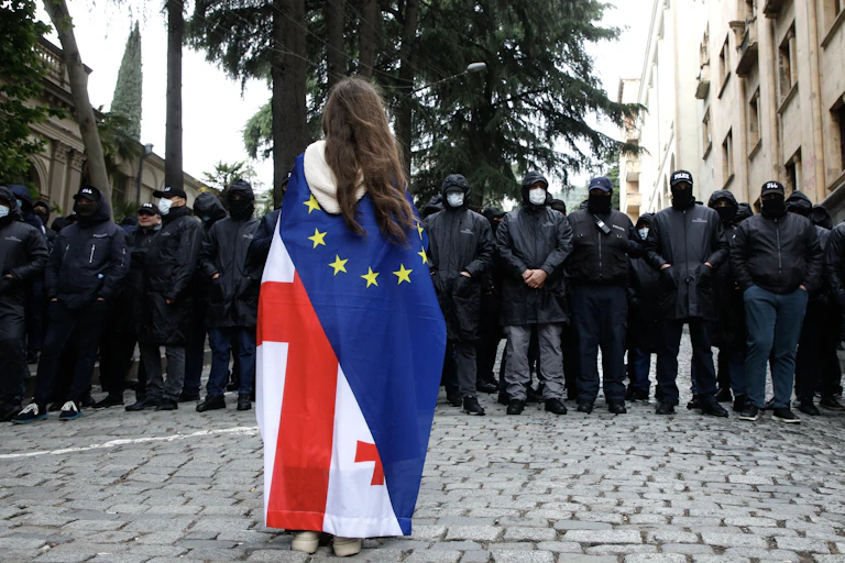A protester wearing a Georgian and European flag faces off with policemen blocking a street.