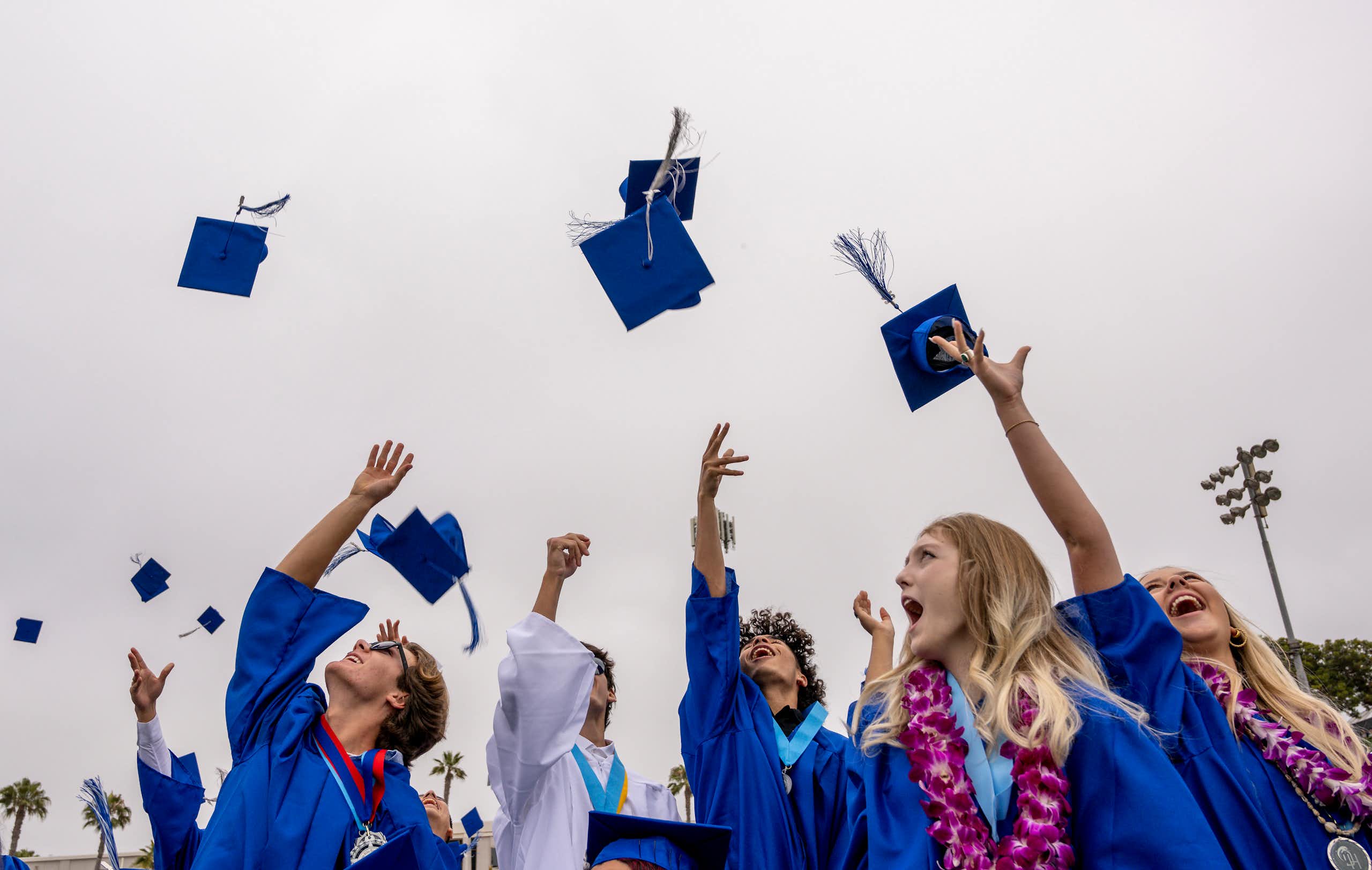 Young men and women wearing blue gowns throw their blue caps in the air in celebration.