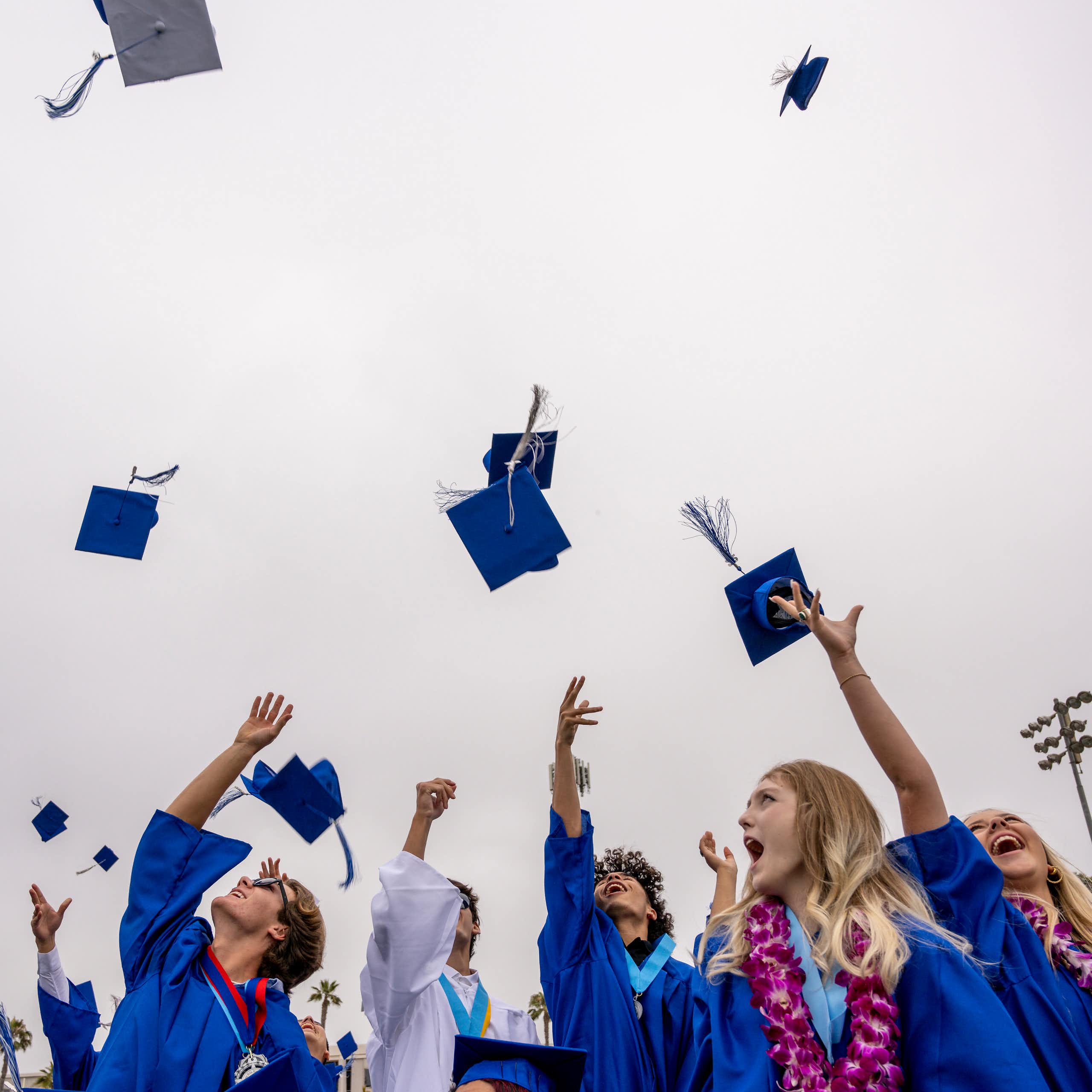 Young men and women wearing blue gowns throw their blue caps in the air in celebration.
