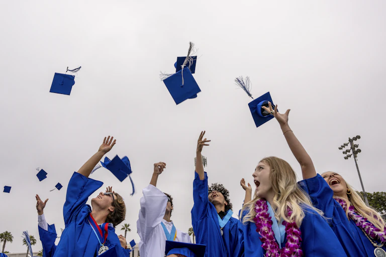 Young men and women wearing blue gowns throw their blue caps in the air in celebration.