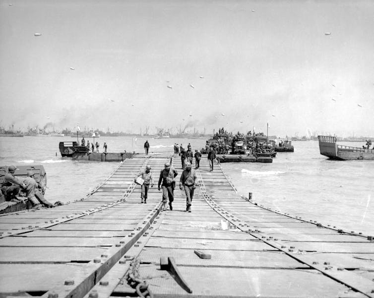 People in uniform walk across a series of attached boxes floating on the water.  Ships can be seen in the background.