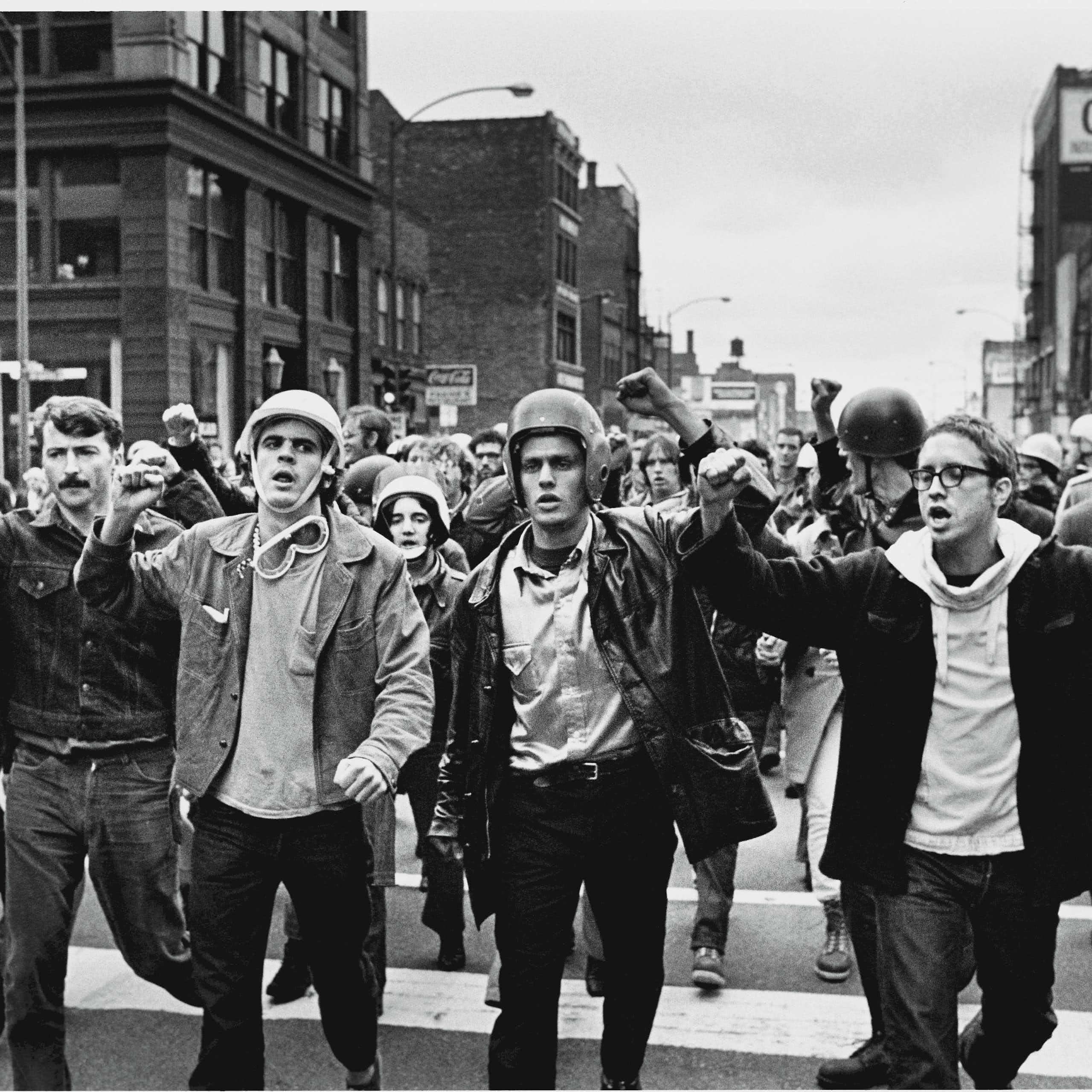 A group of young men, some of them wearing helmets, raise their fists and march down a street in a black and white photo.