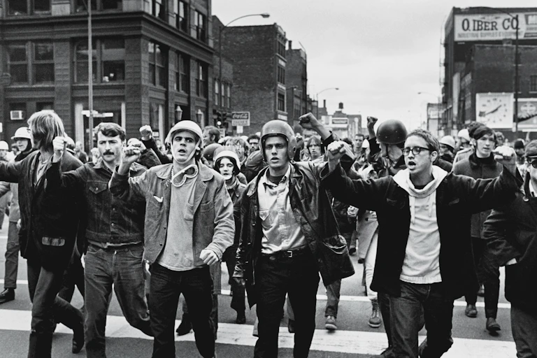 A group of young men, some of them wearing helmets, raise their fists and march down a street in a black and white photo.