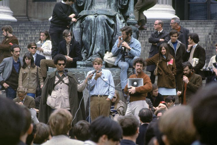 In a vintage-look photo, men and women stand together under a statue.  One of the men speaks into a microphone as people watch.