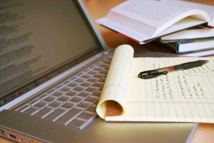 A laptop computer with a yellow legal pad lying on it, next to some books.