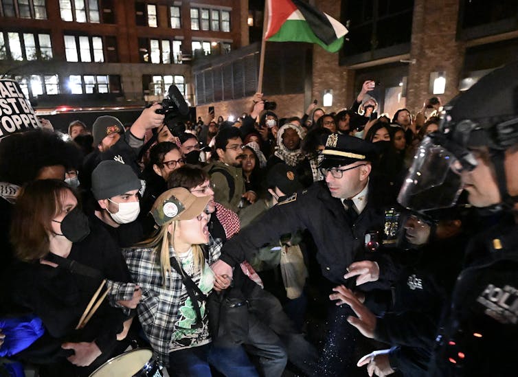A large crowd of people, one of them holding a Palestinian flag, appear to yell and face a line of police officers.