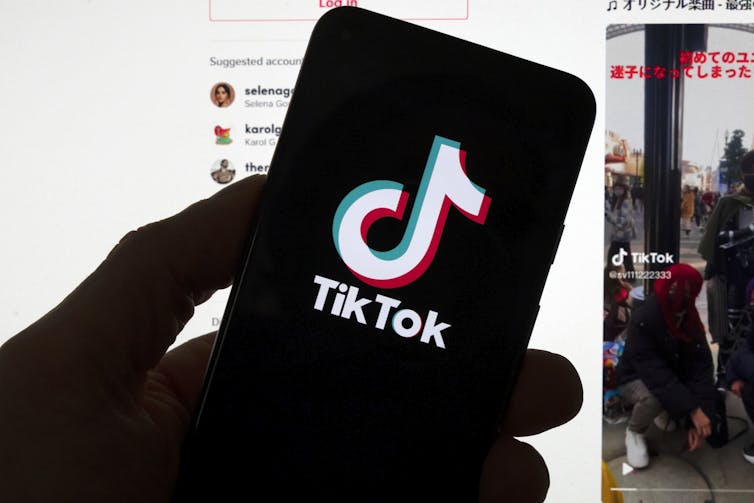 The TikTok logo is seen on a mobile phone in front of a computer screen which displays the TikTok home screen