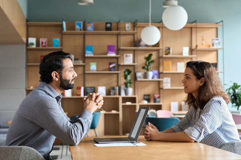A young woman sits across a desk from a middle-aged man. They appear to be having a conversation.