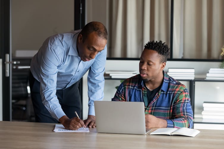 A middle-aged man leans over to write something on a piece of paper sitting on a desk. Beside him, a younger man sits behind a laptop, listening to him speak.