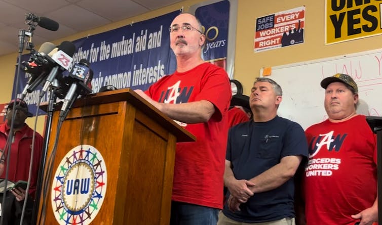 Man stands at a podium looking sad, flanked by other men looking shocked.