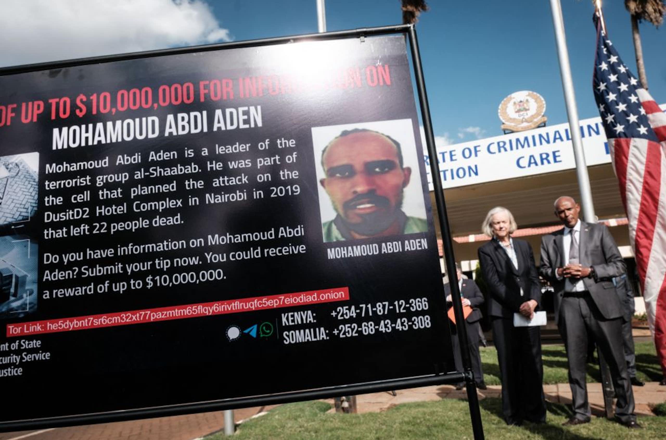 A man and woman standing in the background of a black poster announcing an award of up to US dollars 10 million for information on the leader of the terrorist group al-Shabaab