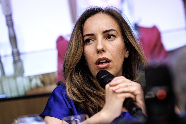 A woman with long brown hair speaks into a handheld microphone