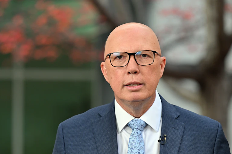 A bald man with black rimmed glasses stands in front of a tree with red leaves
