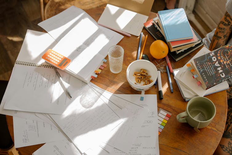 A table is crammed with papers, textbooks, mugs, and an orange.