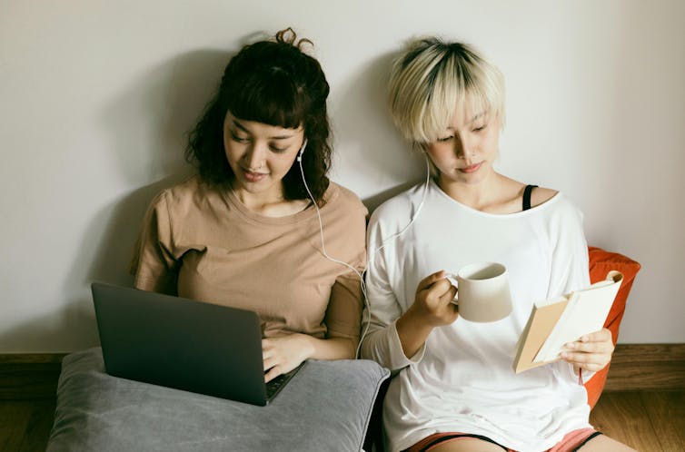 Two young women sit side by side, sharing earphones. One reads a book, one works on a laptop.