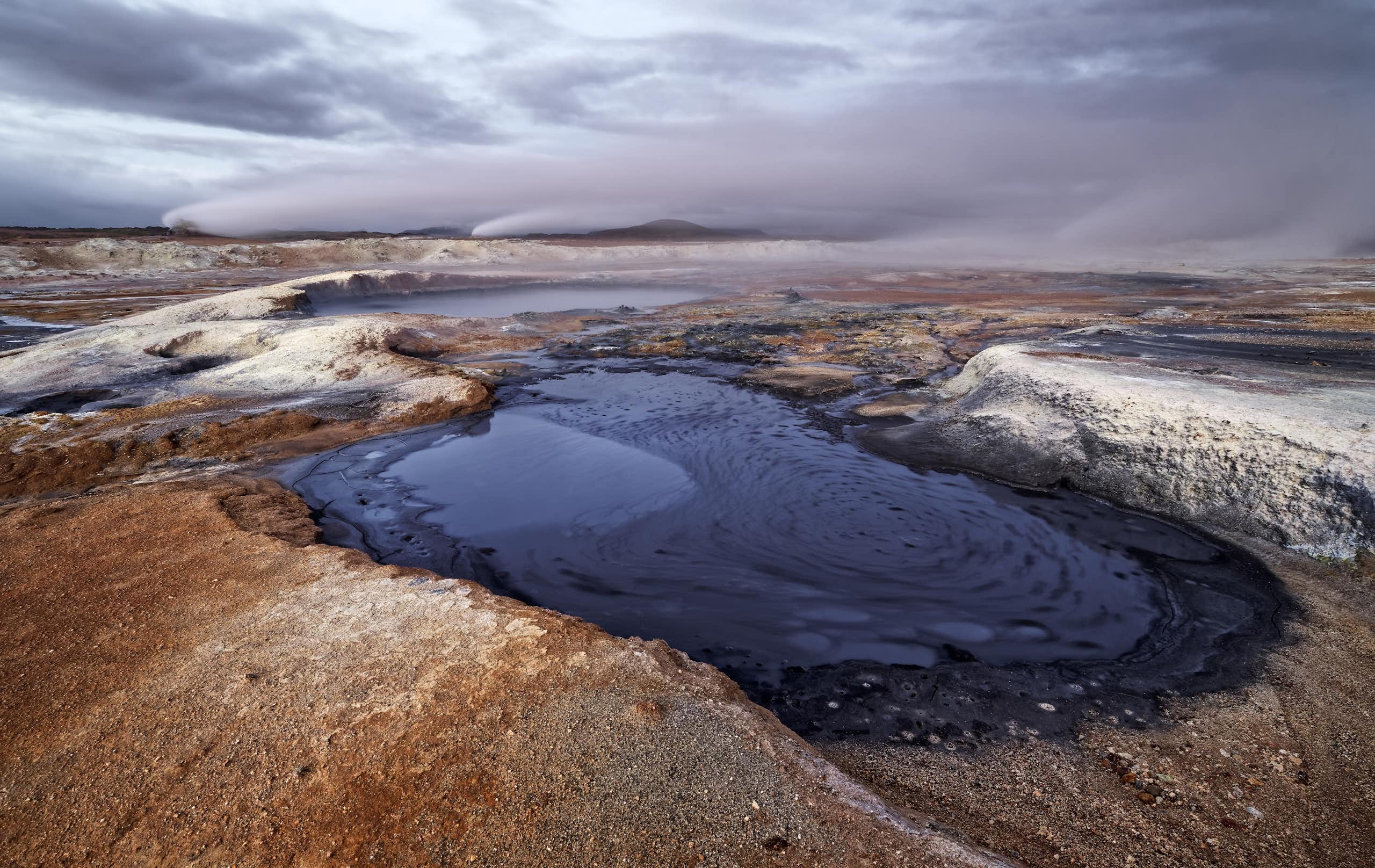 A barren volcanic landscape with a small pool of dark blue water at the forefront.