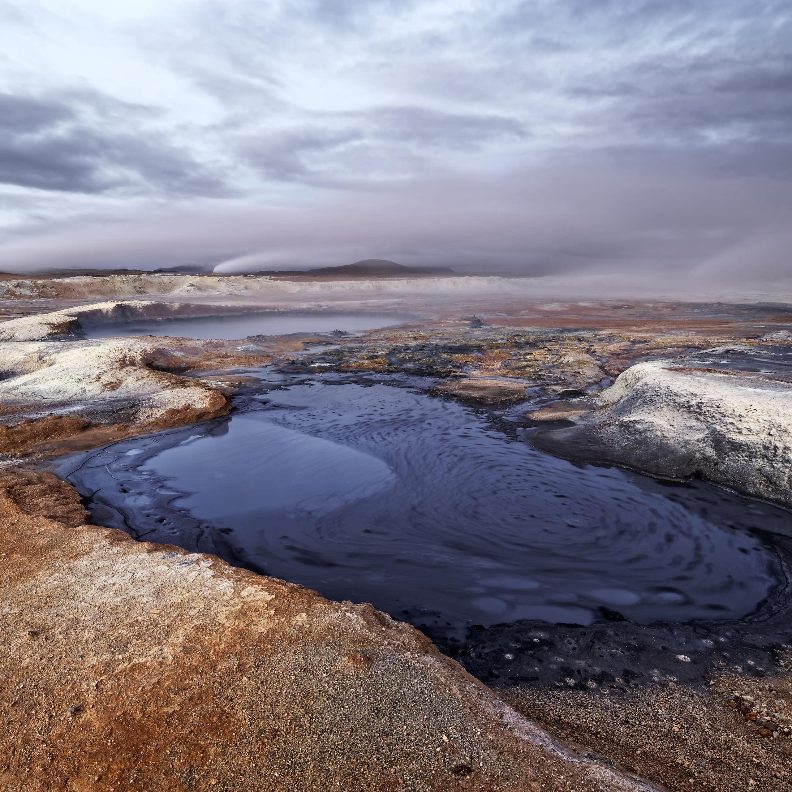 A barren volcanic landscape with a small pool of dark blue water at the forefront.