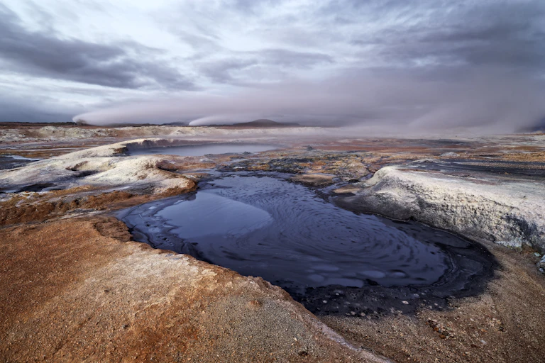 A barren volcanic landscape with a small pool of dark blue water at the forefront.