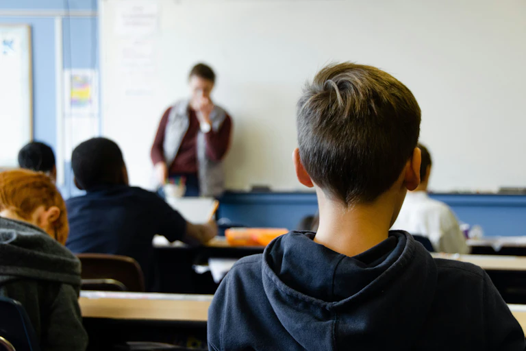 A group of young people sit in a classroom. A teacher speaks at the front of the room in front of a white board.